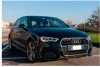 Une Audi noire, garée sur un parking. La voiture est vue de face avec un léger angle, montrant son côté avant droit. L'arrière-plan montre un ciel clair et des bâtiments au loin.