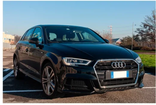 Une Audi noire, garée sur un parking. La voiture est vue de face avec un léger angle, montrant son côté avant droit. L'arrière-plan montre un ciel clair et des bâtiments au loin.